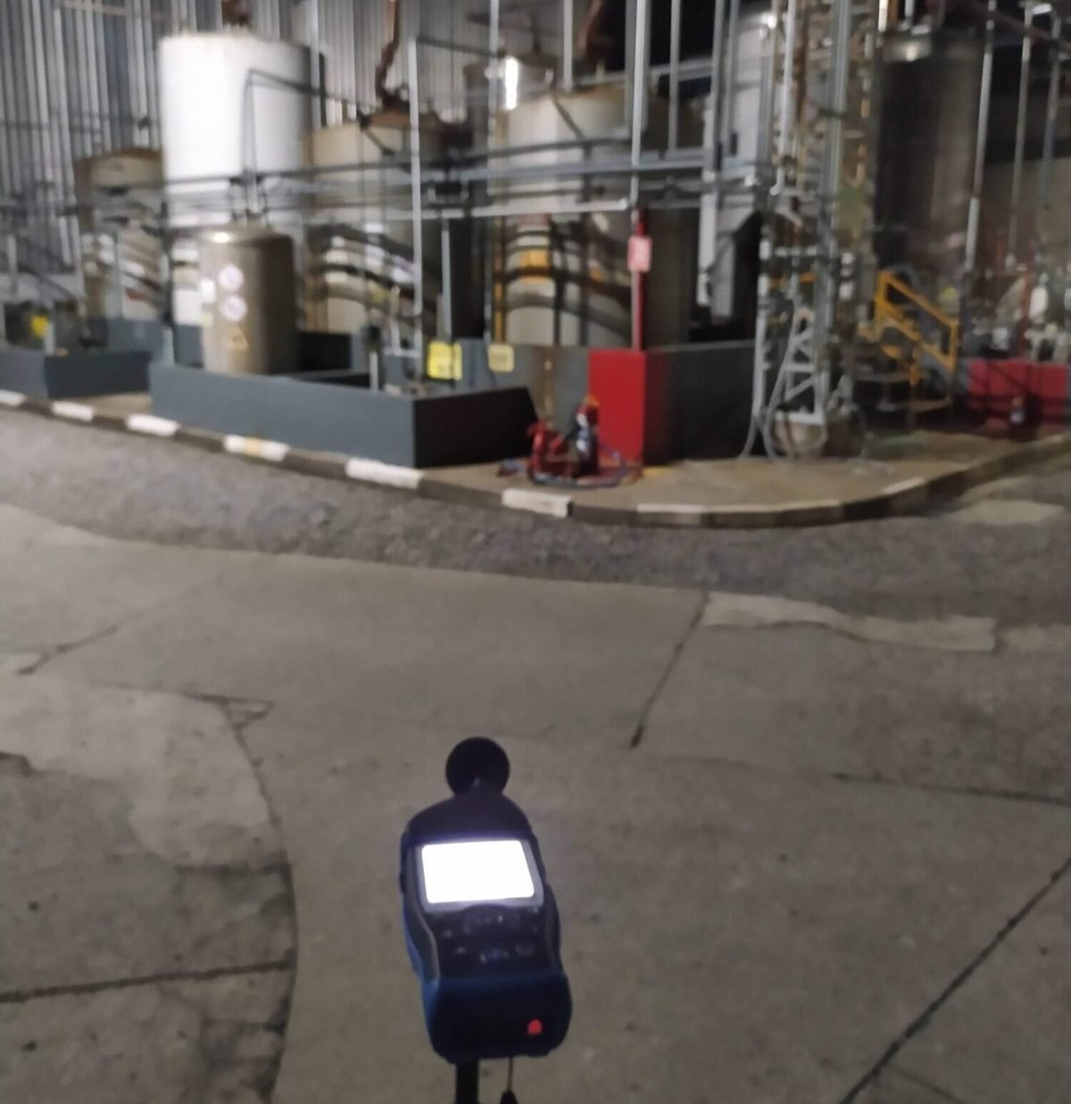 Medição acústica noturna em área industrial com tanques e tubulações ao fundo